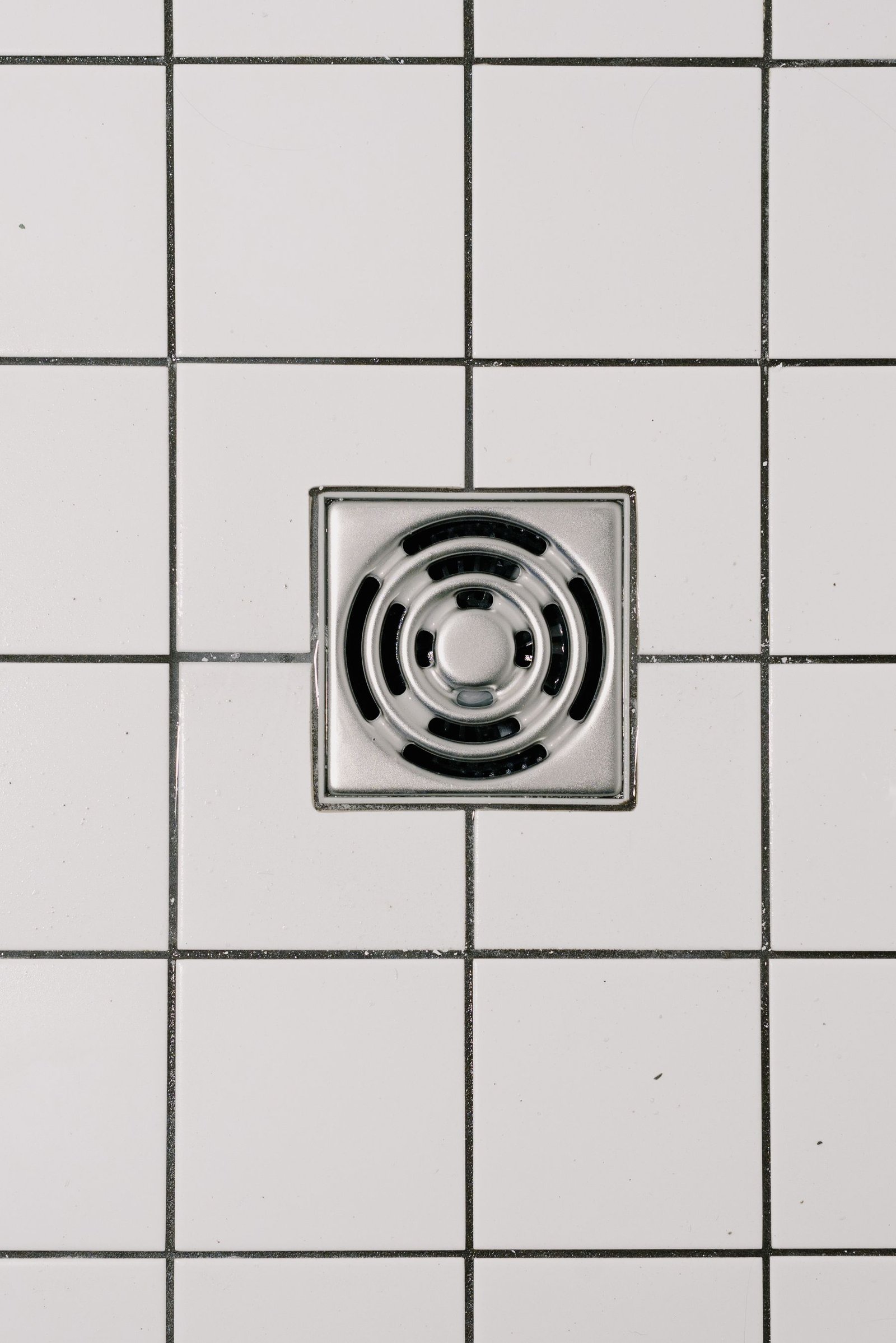 A square metal shower drain set in a white tiled floor with dark grout lines.