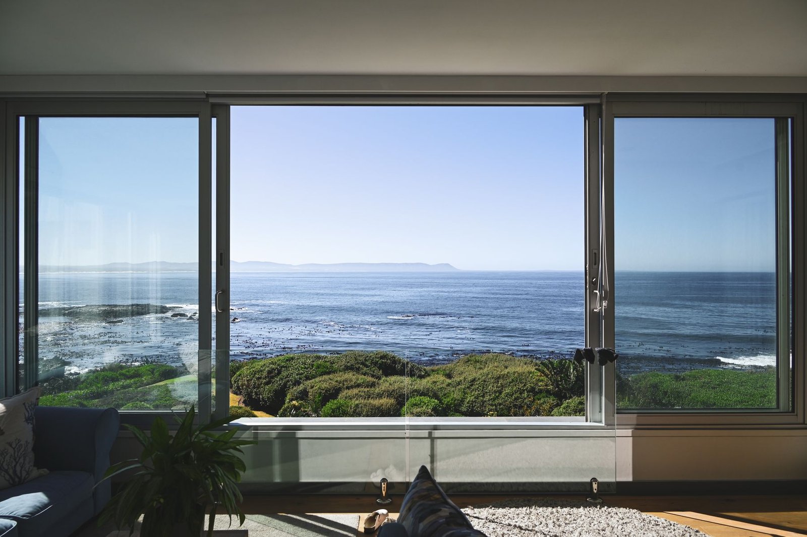 Sunlight enters a living room through broad glazing overlooking a rocky shoreline and the sea.