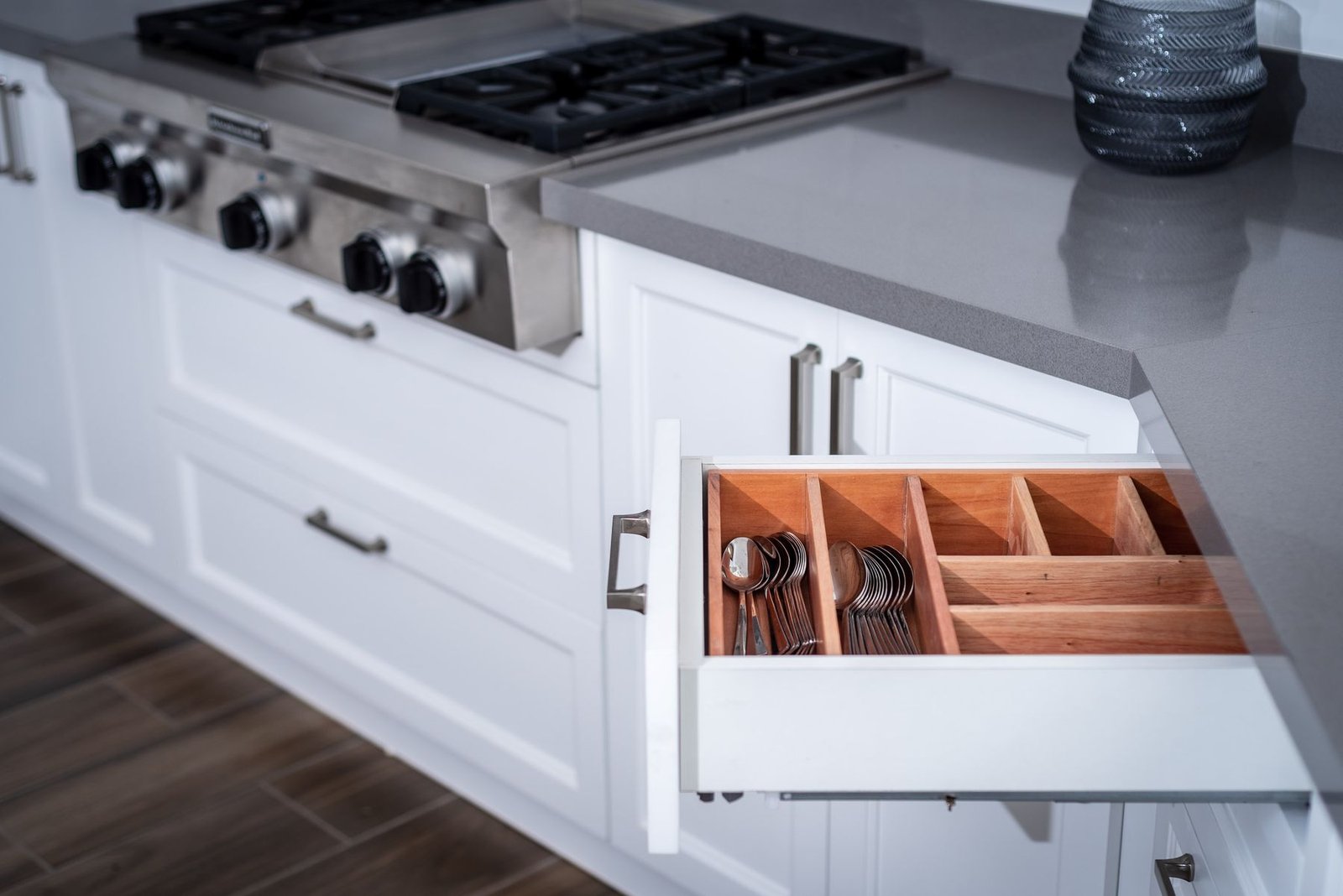 An open kitchen drawer with wooden utensil dividers beside white cabinets and a stainless steel cooker.