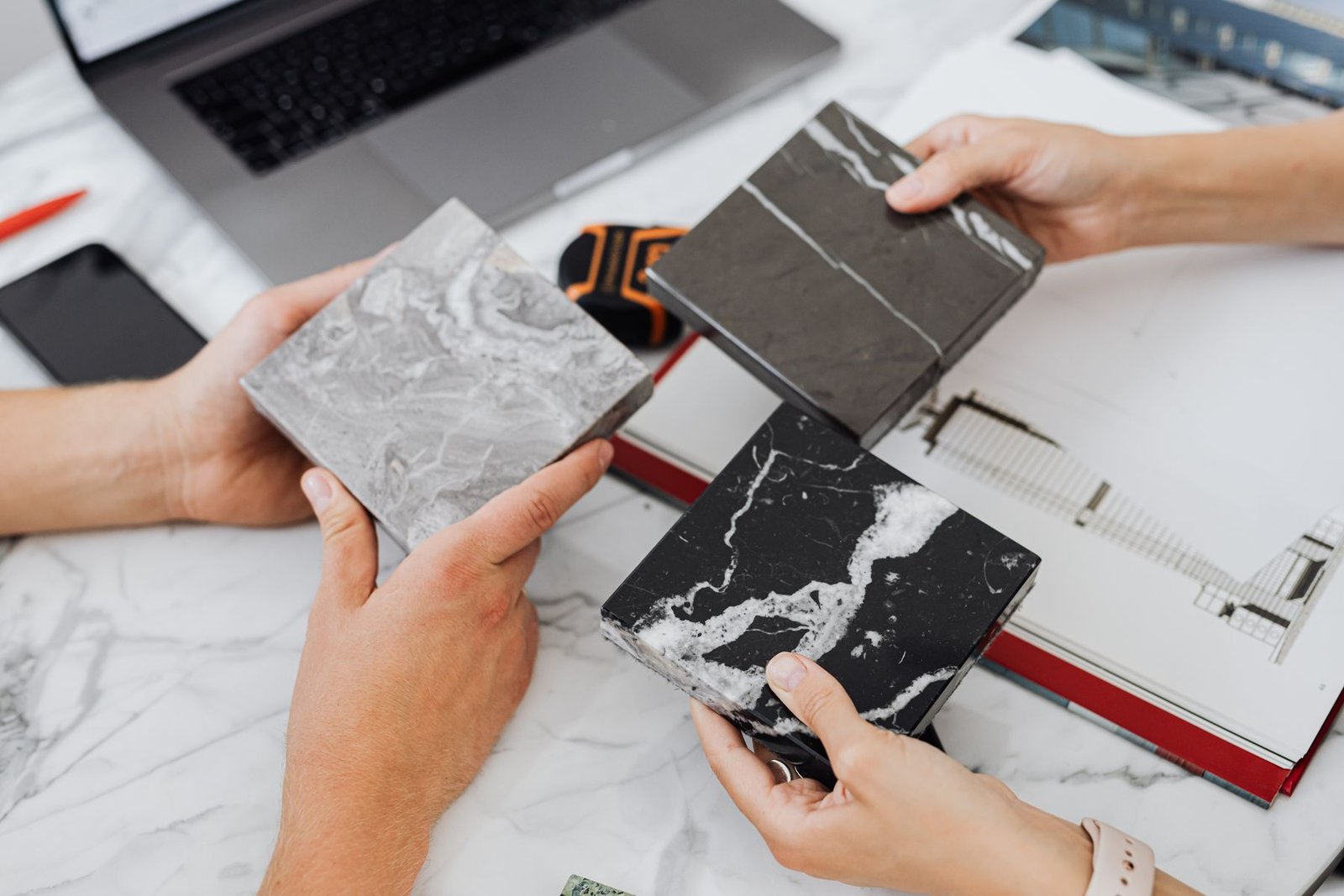 Hands comparing three gray and black veined tile samples beside a laptop and an open design notebook.
